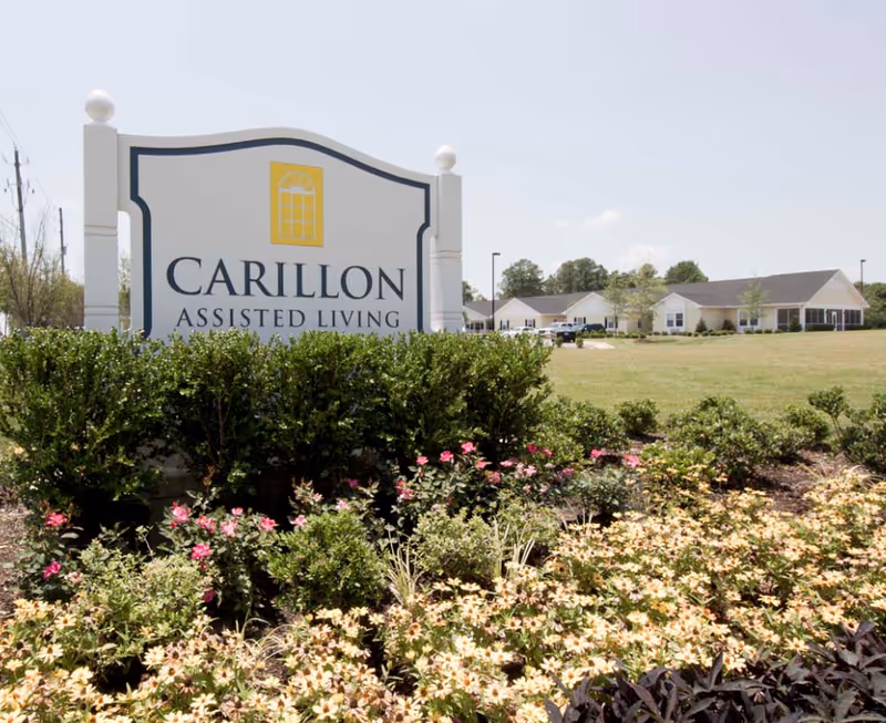 A large landscaped entrance sign reading 'CARILLON ASSISTED LIVING' with flowering shrubs and the facility buildings visible in the background.
