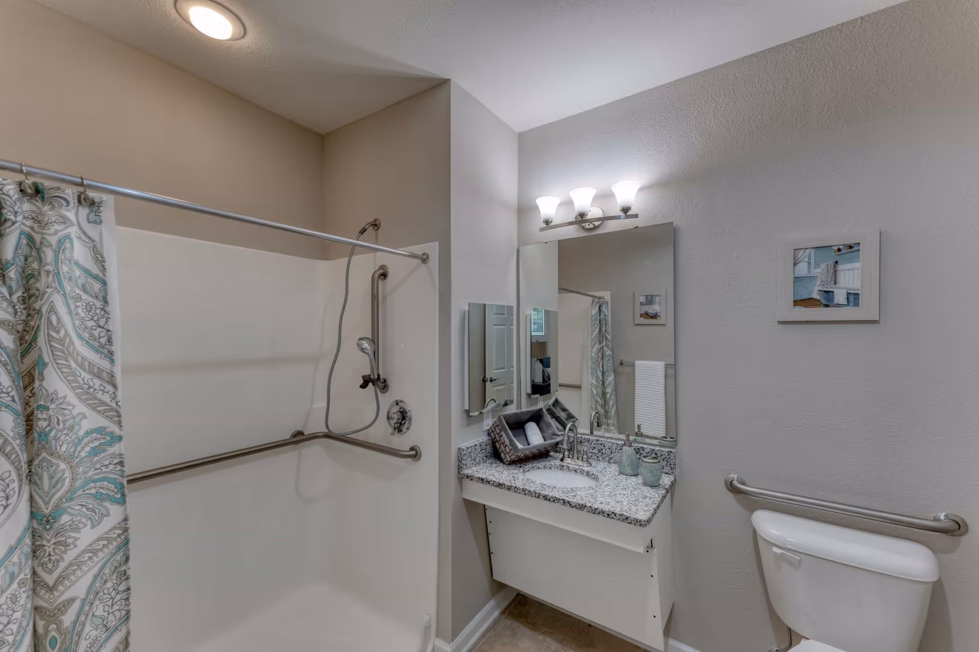 A clean and modern bathroom featuring a shower with a patterned curtain and grab bars, a granite countertop with a sink, a large mirror with three light fixtures above, a small basket with rolled towels, and a toilet with a grab bar on the wall.