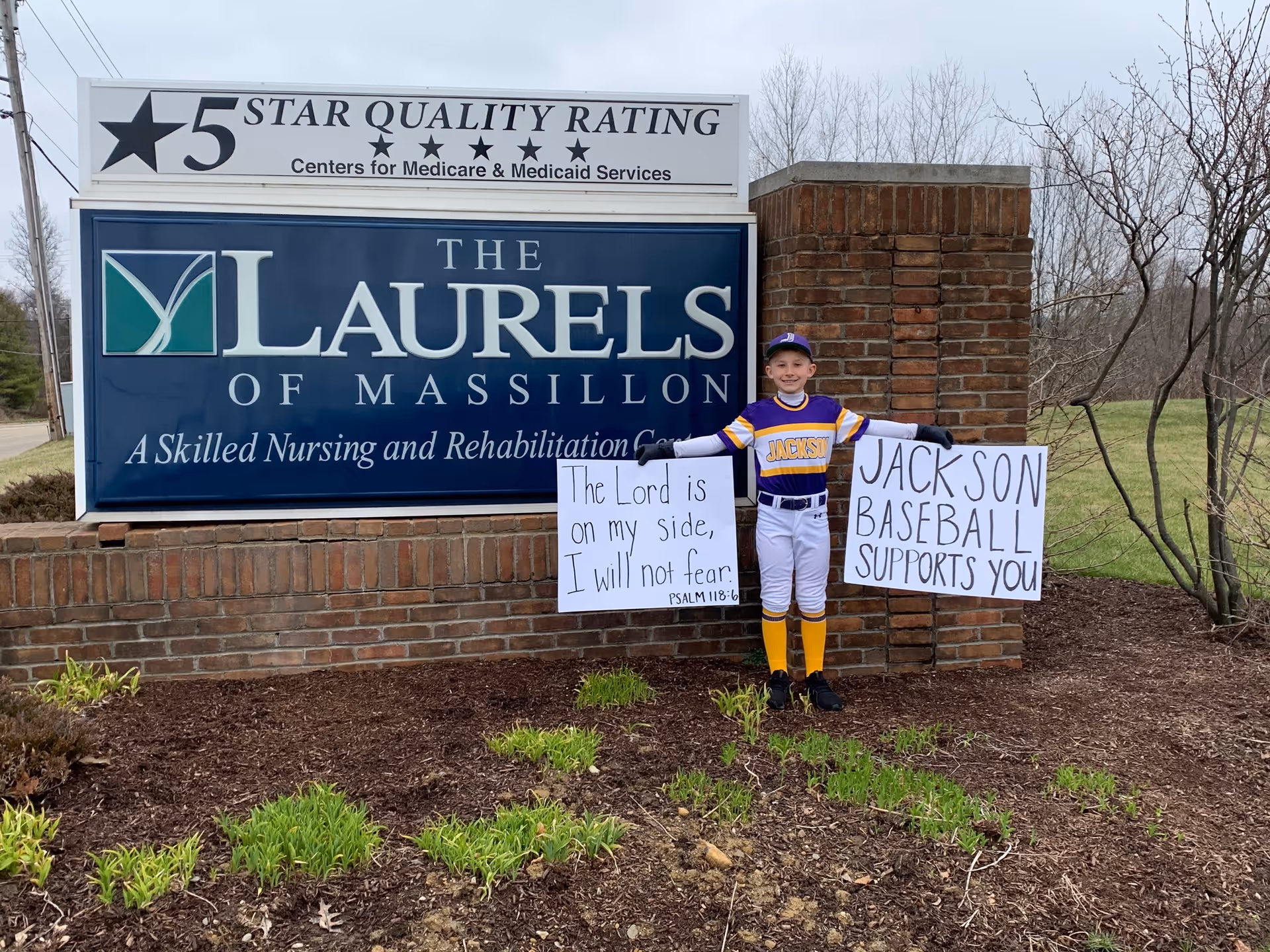 A young baseball player in uniform stands holding supportive signs in front of The Laurels of Massillon facility sign.