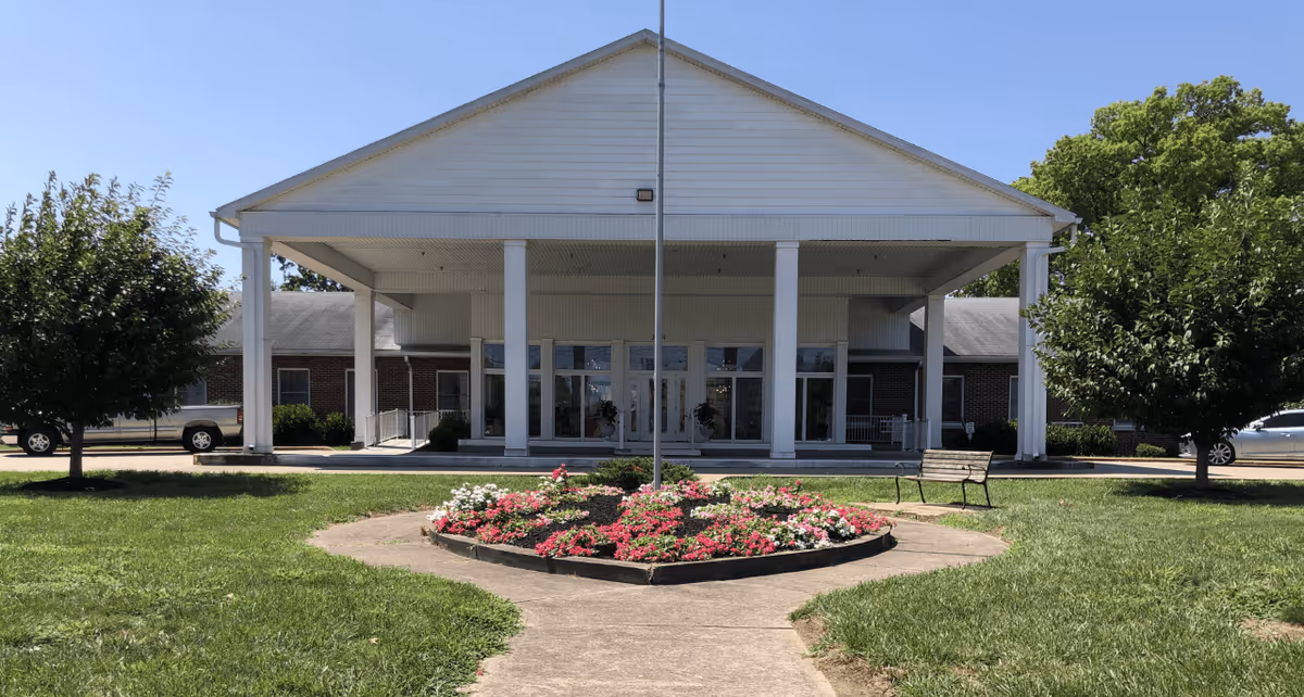 Front exterior view of a single-story building with a large white portico supported by columns. There is a circular flower bed with pink and white flowers in the foreground, flanked by green grass and two trees. A bench is visible near the flower bed on the right side. The sky is clear and blue.