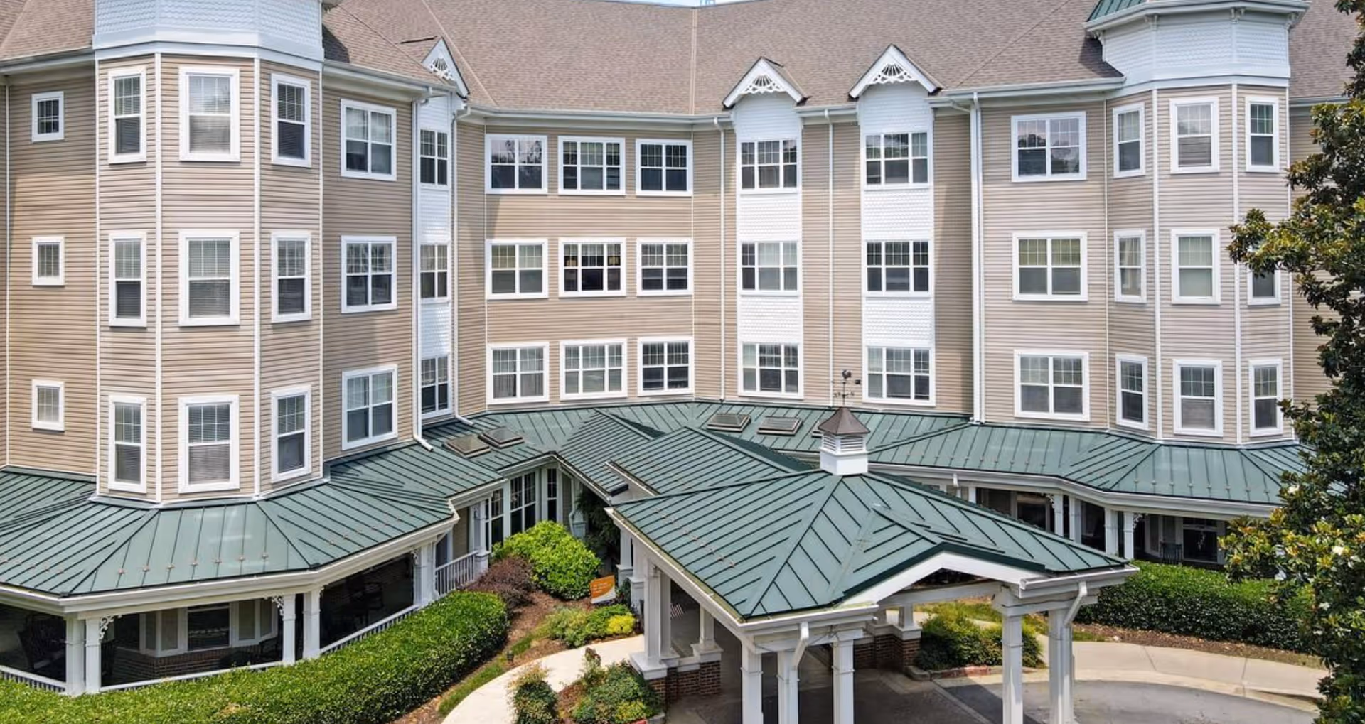 Front exterior of a multi-story beige senior living building with many windows, green metal roofs and a covered porte-cochere surrounded by landscaping.