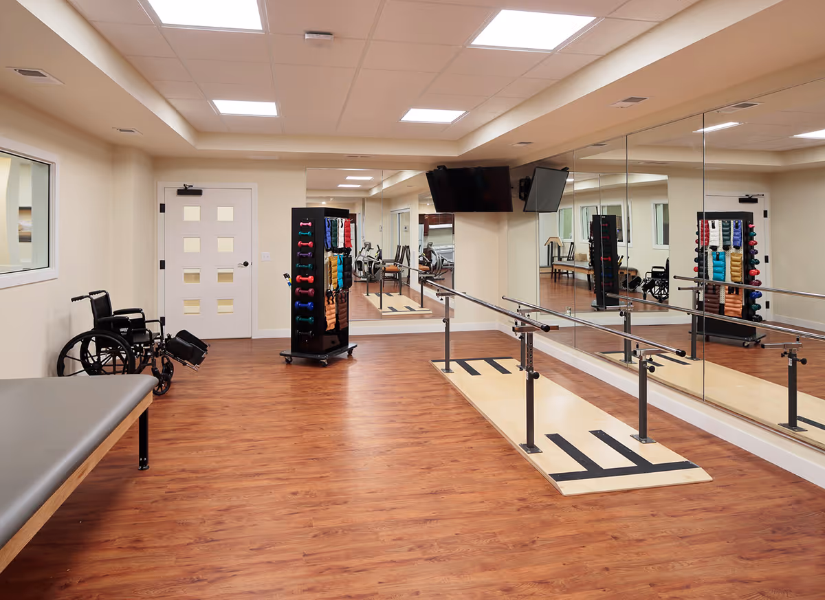 A rehabilitation or physical therapy room with wooden flooring, a wheelchair, a padded therapy table, a rack holding various colored dumbbells and exercise bands, parallel bars for walking exercises, and large mirrors on the wall. Two flat-screen TVs are mounted on the wall above the mirrors.