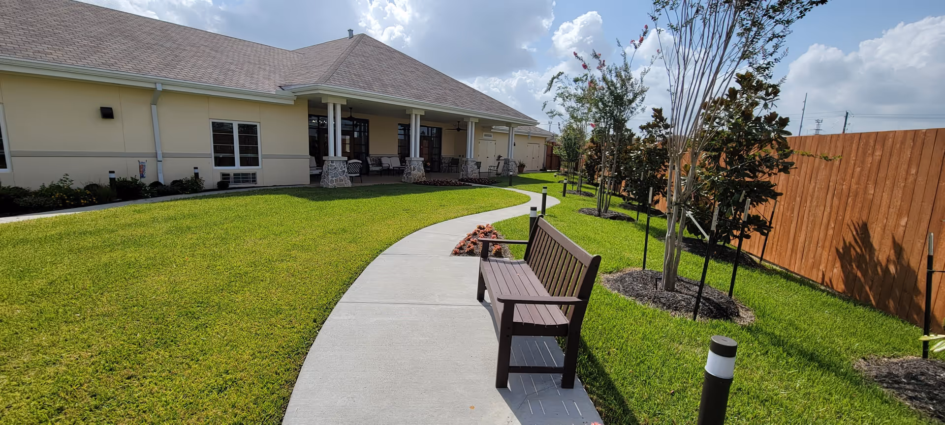 Outdoor area of The Harbor Assisted Living featuring a curved concrete walkway, a wooden bench, green grass, small trees, and a building with a covered patio and seating.