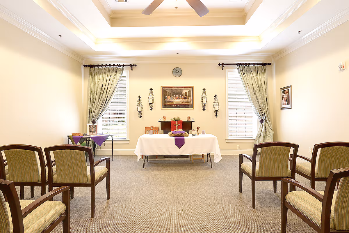 Small multipurpose room with chairs arranged facing a central table/altar beneath a clock and framed artwork.