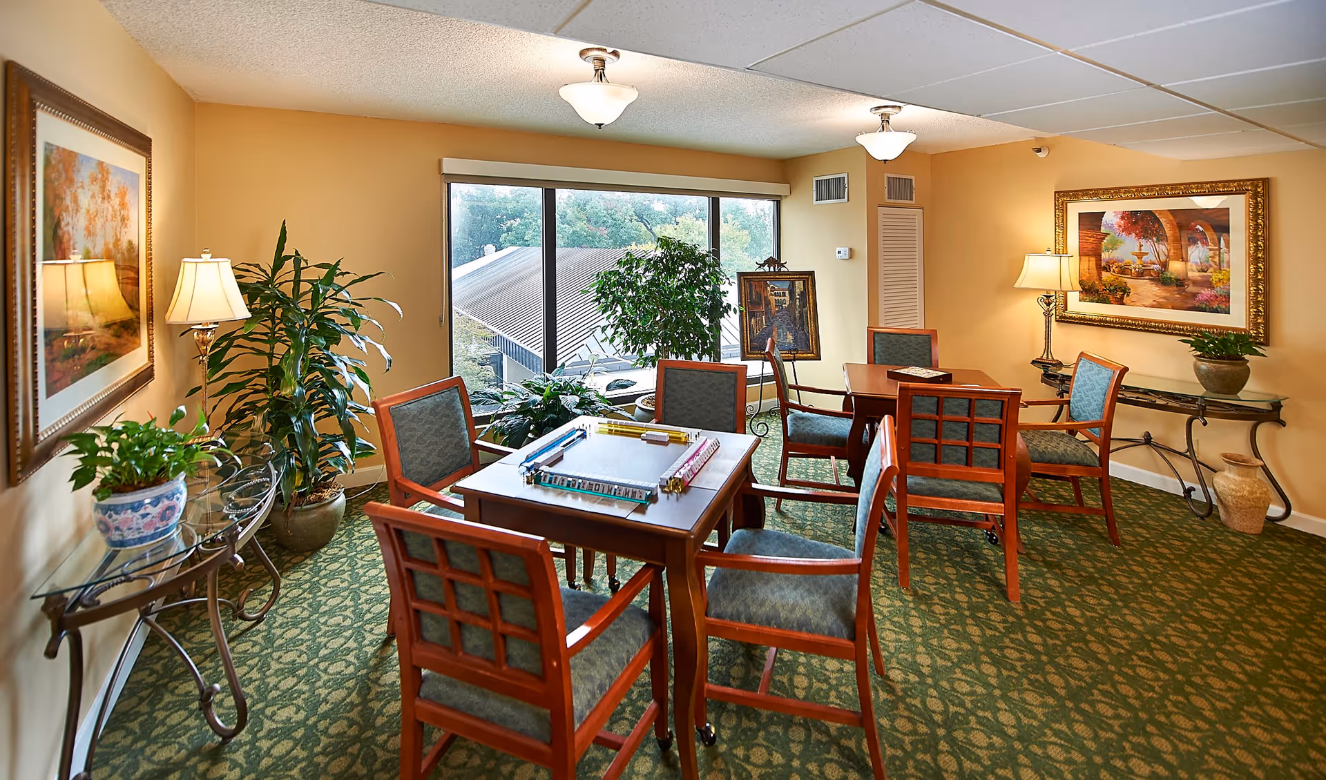A well-lit common room with two wooden tables and several chairs arranged around them. One table has a board game set up. The room features green patterned carpet, beige walls, large windows with a view of trees and a roof outside, framed artwork on the walls, potted plants, and table lamps providing warm lighting.