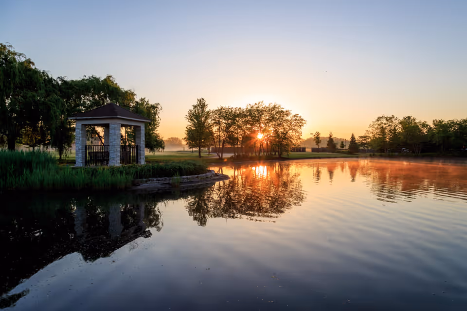 A small lakeside gazebo and trees reflected in a calm pond at sunrise.