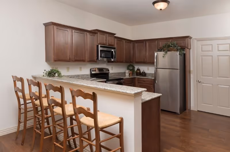 A kitchen with wooden cabinets, granite countertops, stainless steel appliances including a refrigerator, stove, and microwave. There is a breakfast bar with four wooden chairs with woven seats. The floor is wooden and the walls are painted white.