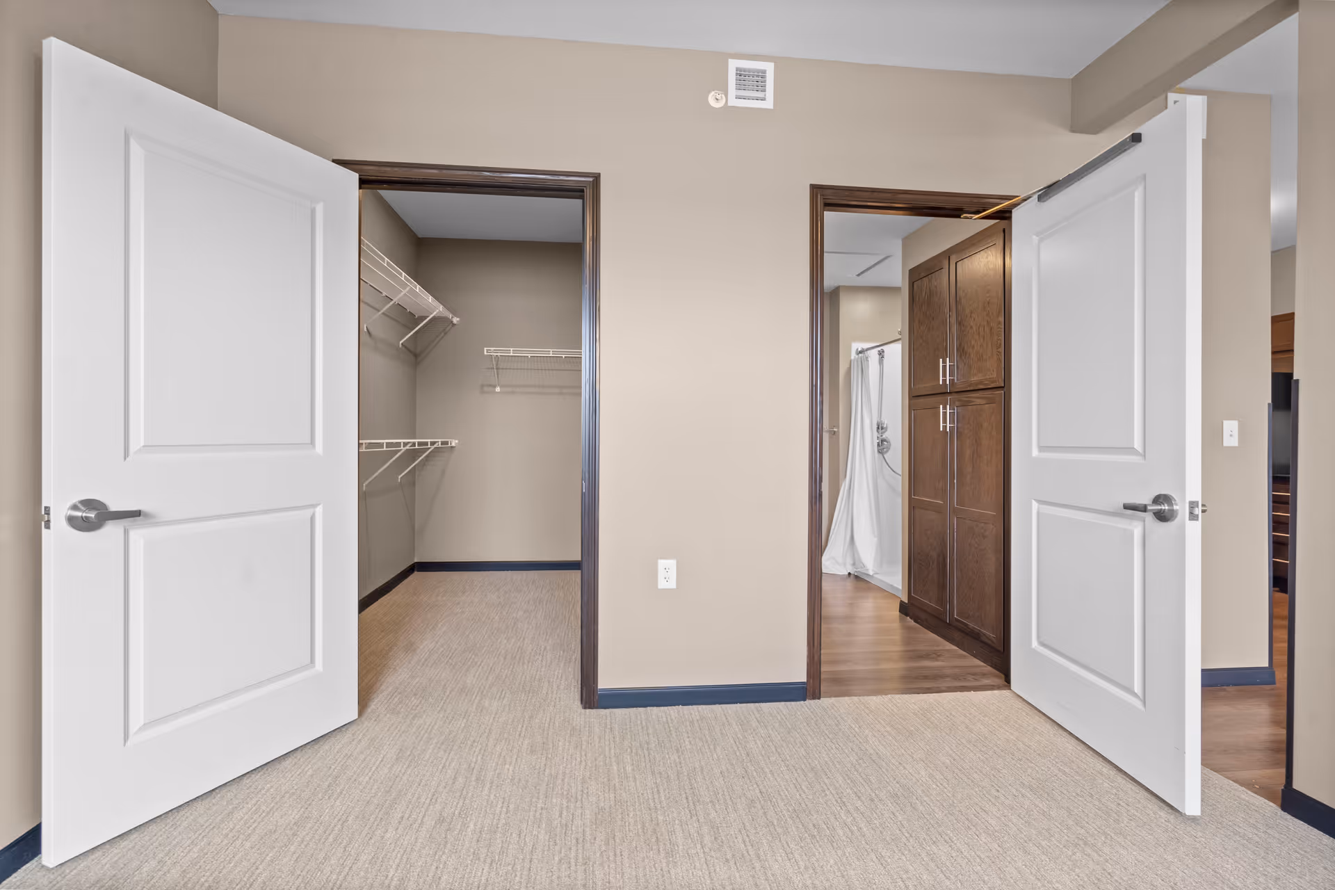 Empty bedroom area with open double doors revealing a walk-in closet on the left and a bathroom with shower and cabinets on the right.