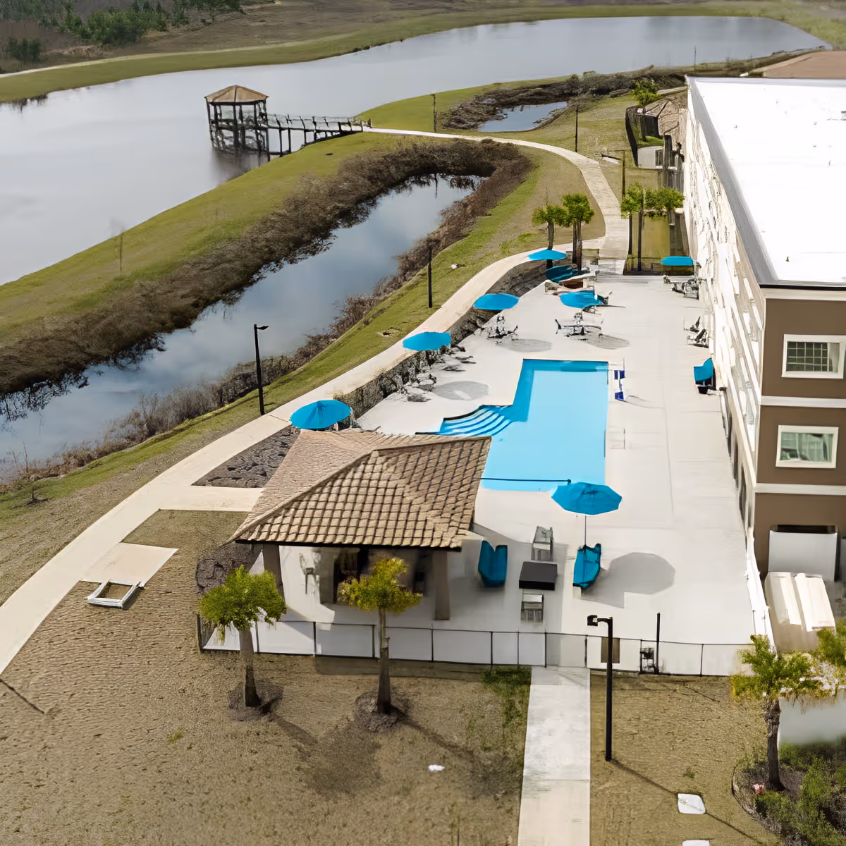 Aerial view of an outdoor pool area next to a multi-story building with several blue umbrellas and lounge chairs around the pool. There is a small covered pavilion with a tiled roof near the pool. The area is surrounded by a walking path, grassy land, and a body of water with a wooden gazebo on a pier extending into the water.