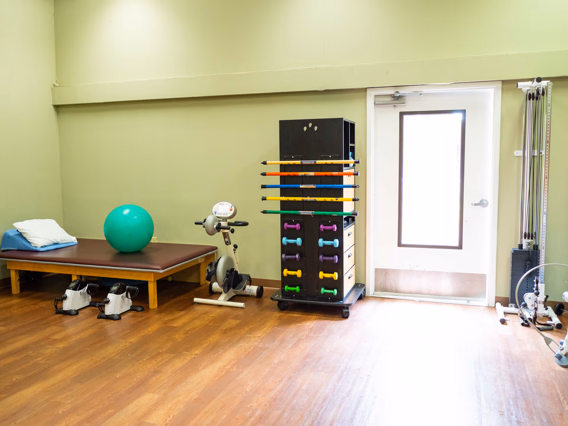 A small exercise room with a padded bench holding a pillow and a green exercise ball, a stationary pedal exerciser, a rack with colorful dumbbells and resistance bars, and a cable pulley machine near a door with a window.