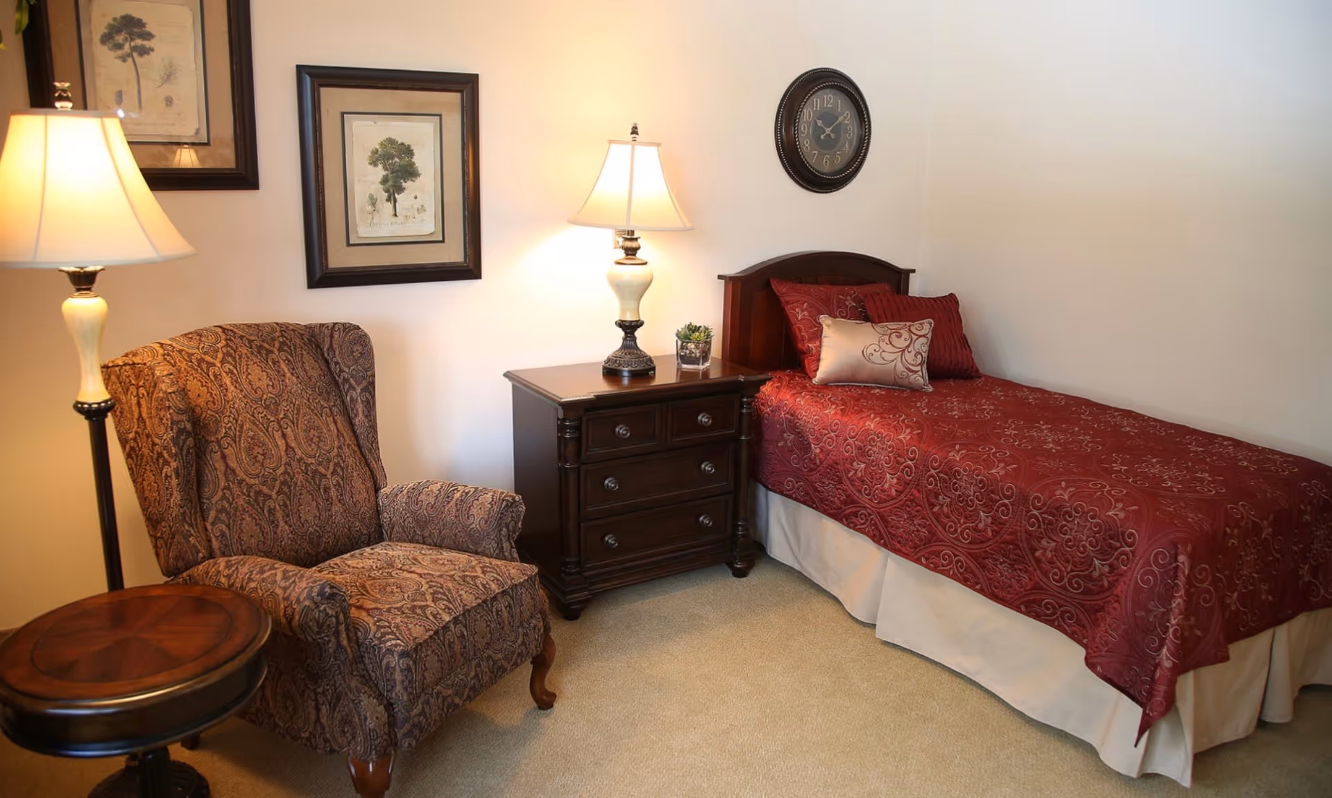 Cozy single bedroom with a red patterned bed, wooden nightstand, upholstered armchair, lamps, and wall art.