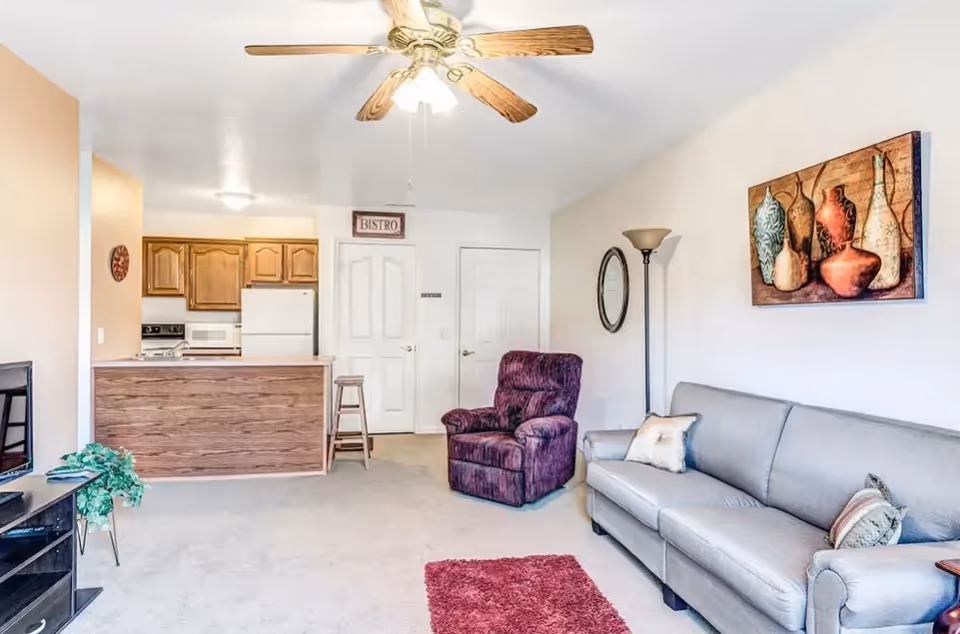 A living room area with a gray couch, a purple armchair, a floor lamp, and a wall painting of vases. The room has a ceiling fan with lights, a small red rug on the carpeted floor, and a kitchen area with wooden cabinets and white appliances in the background.