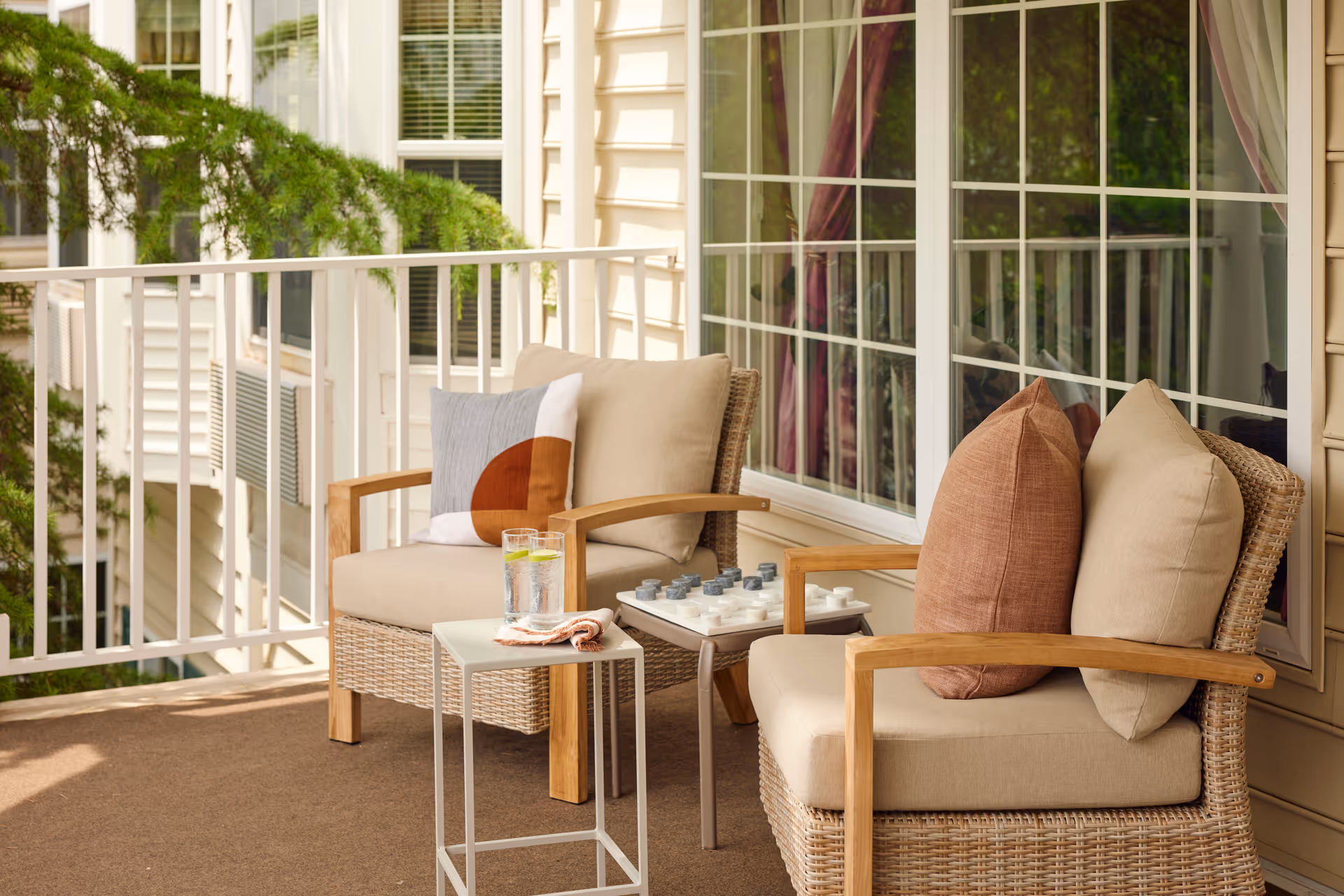 A cozy outdoor balcony area with two wicker armchairs featuring beige cushions and decorative pillows. A small white side table holds two glasses of water with lemon slices and a folded napkin. A chessboard is placed on a small table between the chairs. The balcony has white railings and is adjacent to large windows with white frames and beige siding on the building exterior.