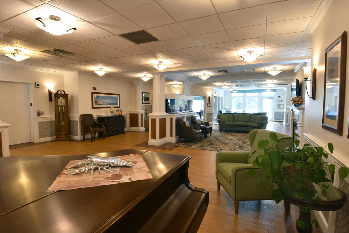 A spacious and well-lit living room area in a senior living facility named Foreside Harbor, featuring green upholstered chairs and sofas, a wooden piano with a decorative silver lobster on top, a large aquarium, a grandfather clock, framed artwork on the walls, and a patterned rug on the wooden floor.