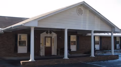 Front exterior view of a single-story brick building with a covered porch supported by white columns. The building has a gabled roof with white siding on the gable end and several windows with dark shutters.
