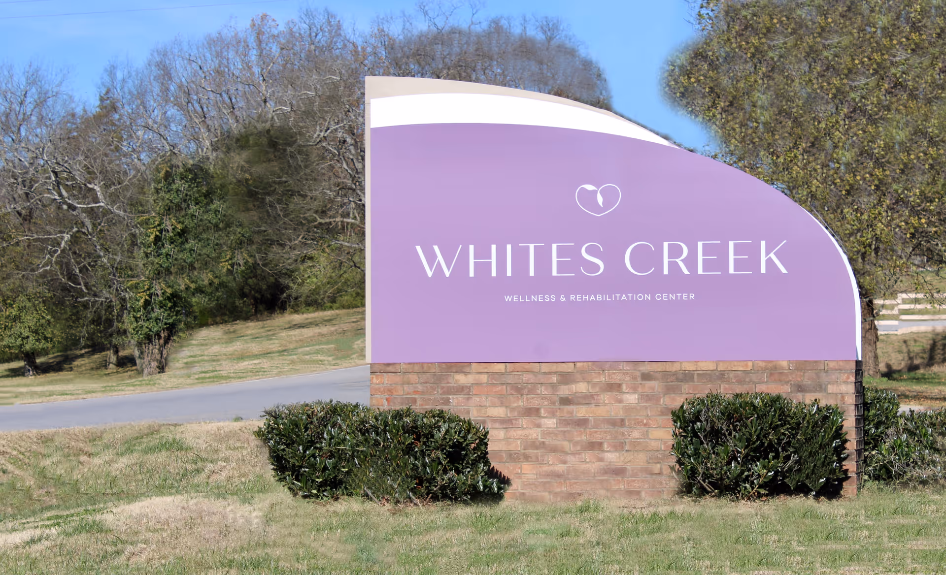 Purple entry sign reading "WHITES CREEK Wellness & Rehabilitation Center" on a brick base with grass and trees behind it.