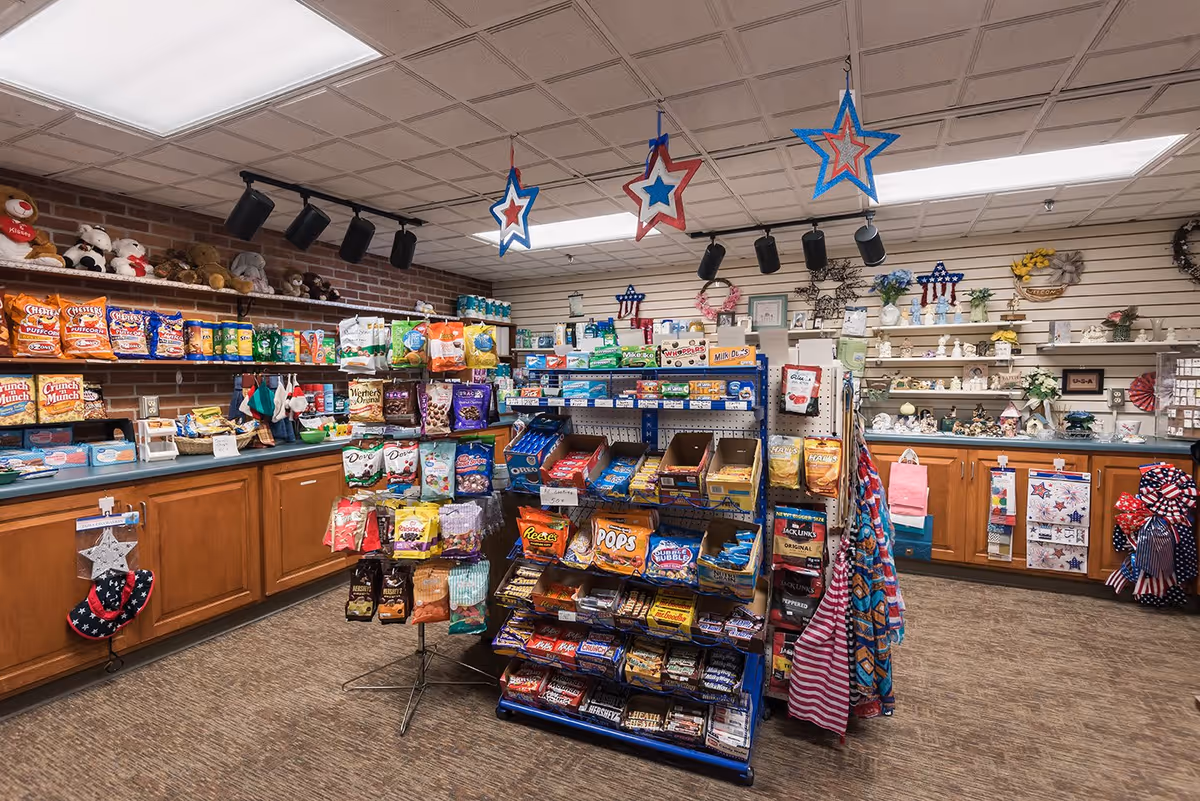 Interior view of a small convenience store or gift shop inside a senior living facility. Shelves and racks are stocked with snacks, candy, and small gift items. The walls have decorative items and shelves with various knick-knacks. The ceiling has hanging star decorations in red, white, and blue colors.
