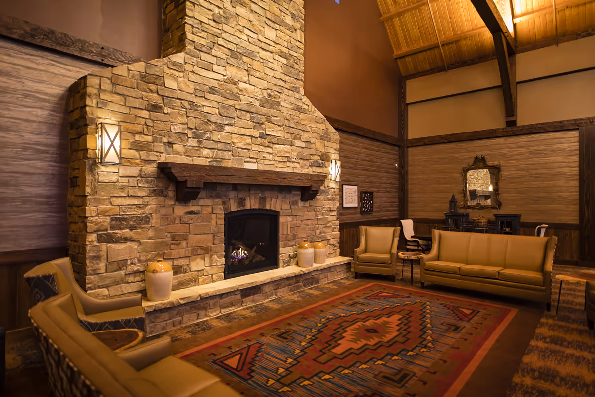 Cozy lounge with a large stone fireplace, sofas and armchairs arranged around a patterned rug under a vaulted wooden ceiling.