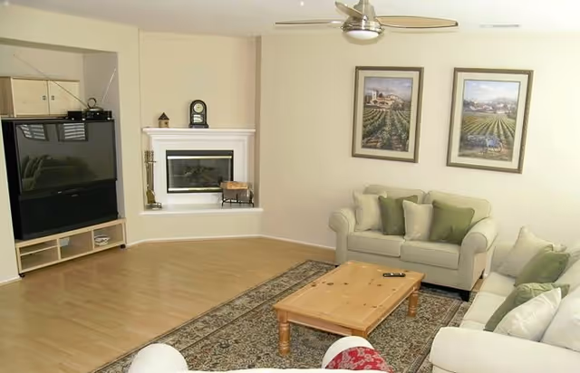 A cozy living room with light-colored walls and wooden flooring. The room features two beige sofas with green and beige cushions, a wooden coffee table with a remote control on it, a patterned area rug, a corner fireplace with a clock and decorative items on the mantel, and a large flat-screen TV on a wooden stand. Two framed landscape paintings hang on the wall above one of the sofas.