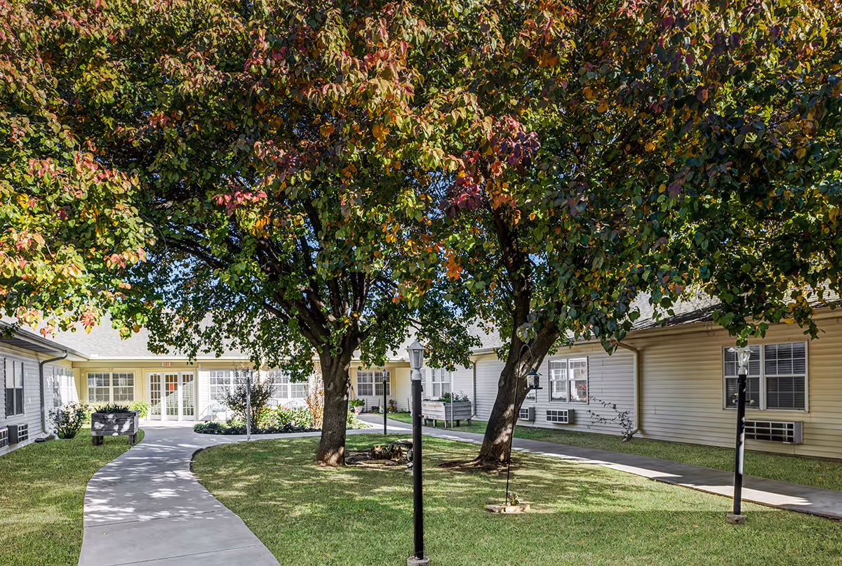 Outdoor courtyard area at Chisholm Place featuring a curved concrete walkway, green grass, two large trees with leaves turning autumn colors, lamp posts, and a swing hanging from one of the trees. The courtyard is surrounded by a single-story building with white siding and multiple windows.