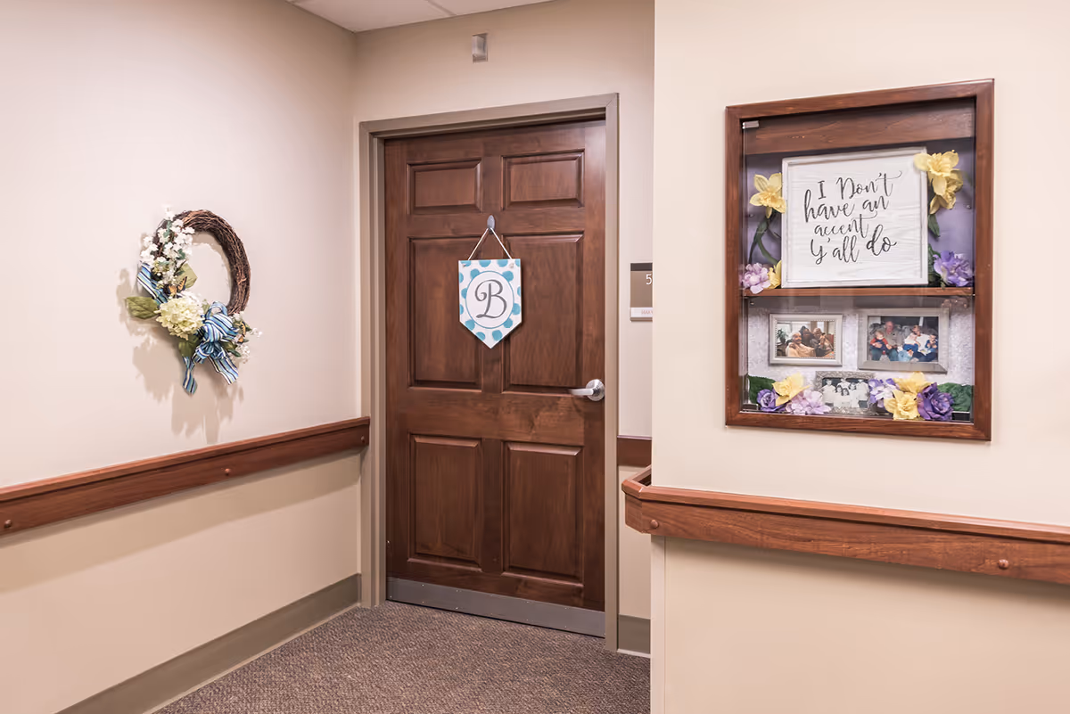 Wooden door in a senior living hallway decorated with a hanging monogram sign, wall wreath, and a framed bulletin case with photos and flowers.