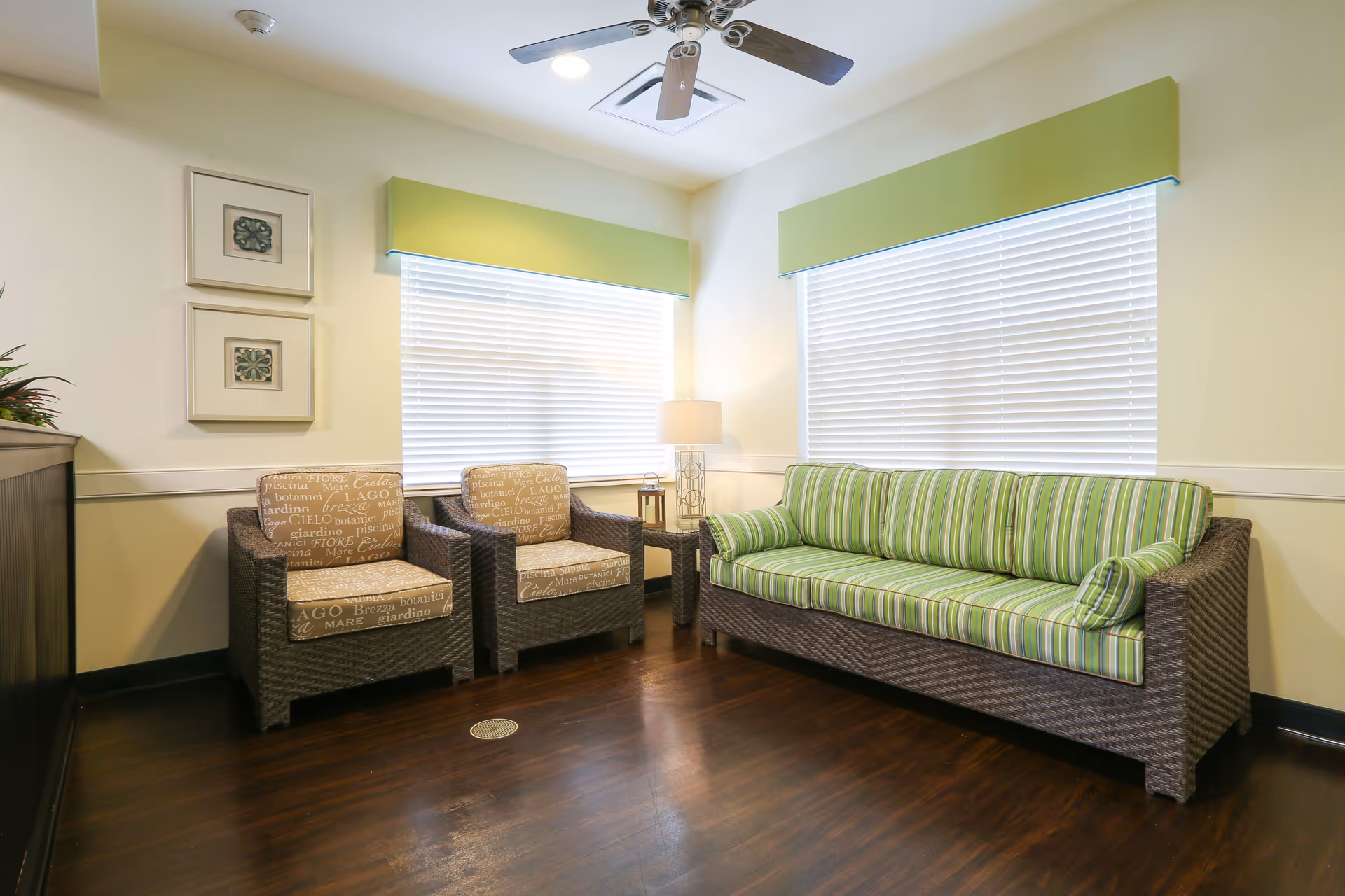 A cozy sitting area in a senior living facility with two wicker armchairs featuring beige cushions with text patterns, and a wicker sofa with green striped cushions. The room has two large windows with white blinds and green valances, a wooden floor, a ceiling fan with lights, a small side table with a lamp, and framed artwork on the wall.