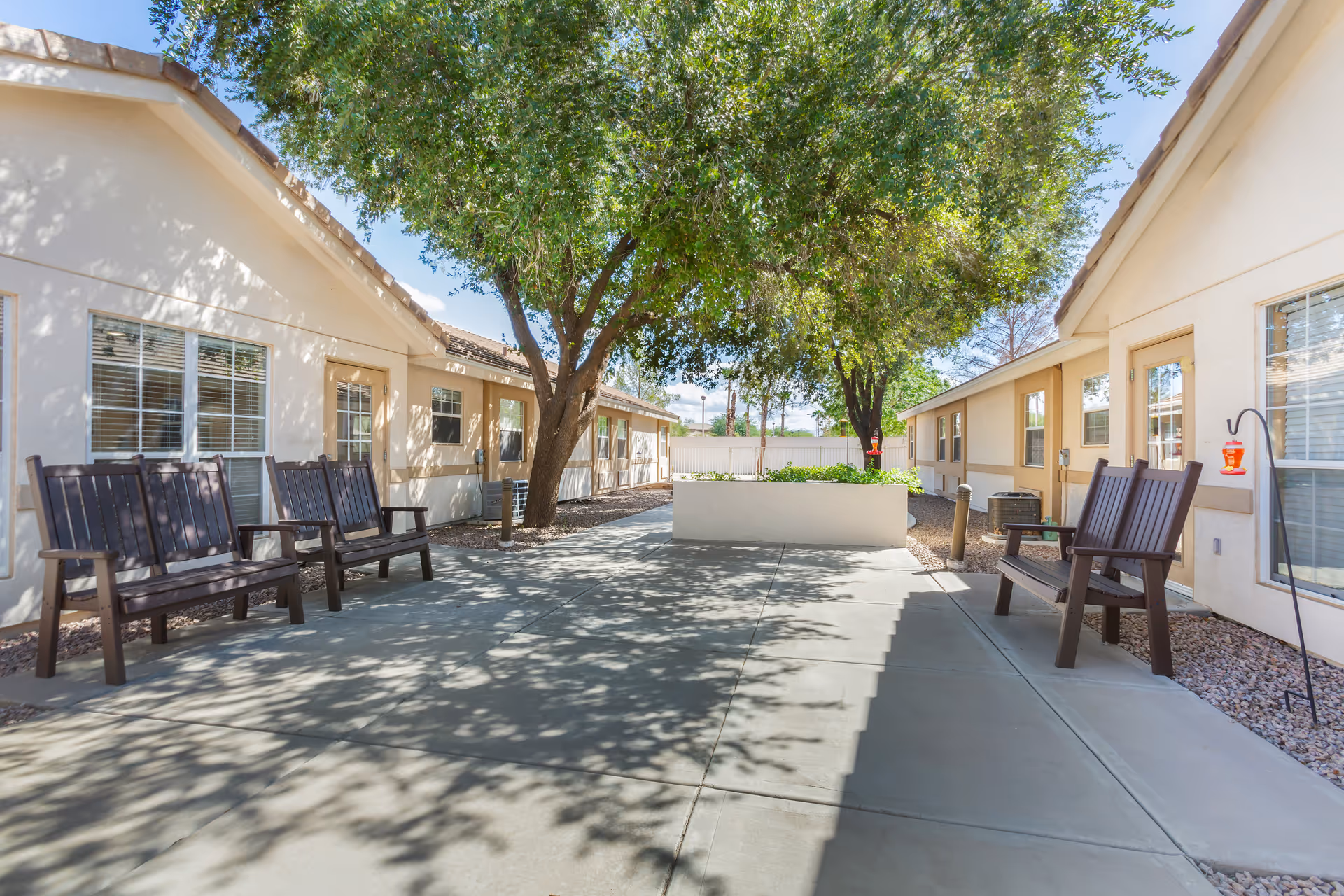 Sunlit courtyard between single-story residential buildings with benches, trees, and a central planter.