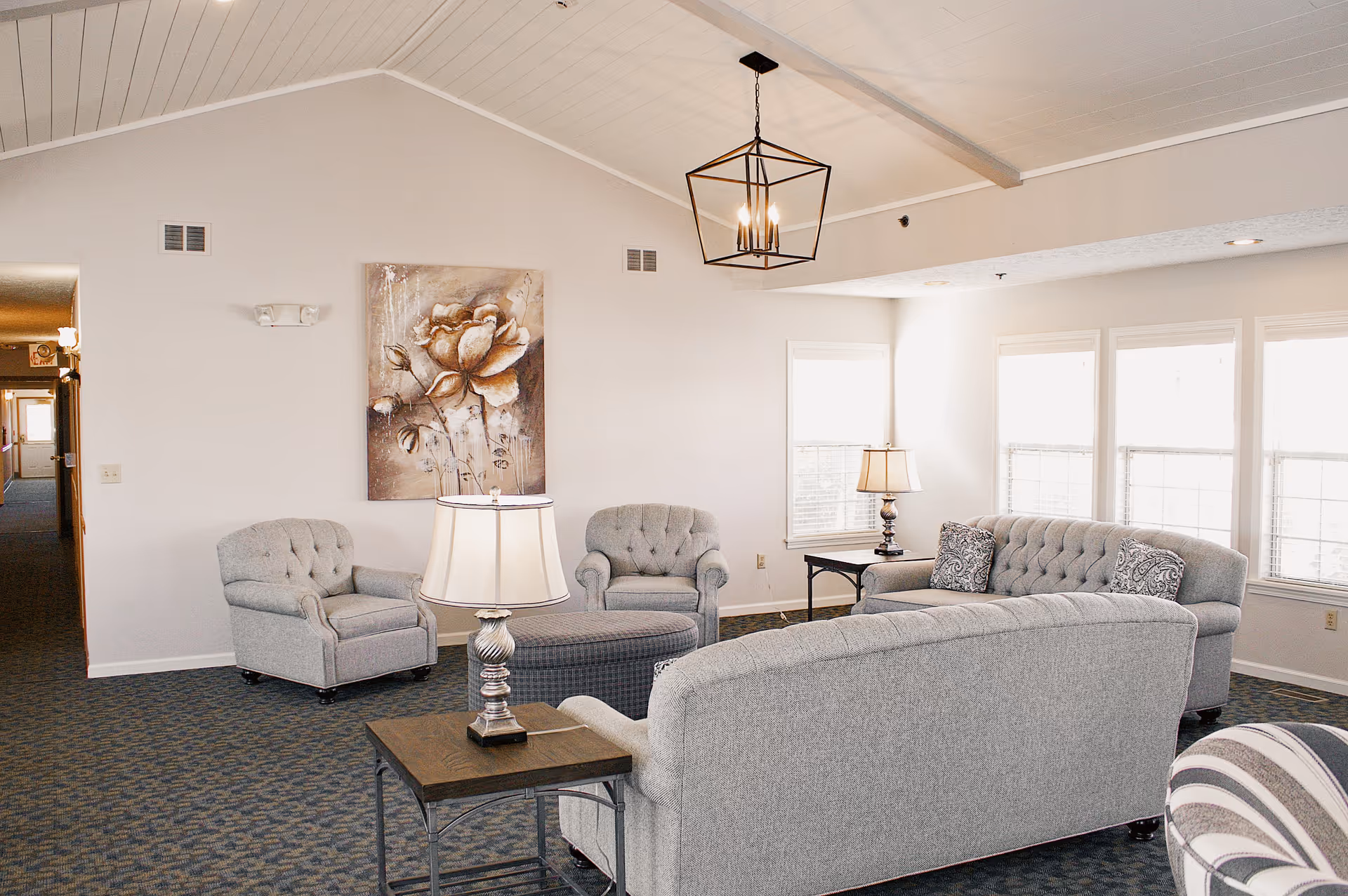 A bright living room area in a senior community featuring gray upholstered sofas and armchairs arranged around a central ottoman. There are two table lamps on side tables, a large floral painting on the wall, and multiple windows letting in natural light. The ceiling is vaulted with white paneling and a modern geometric chandelier.
