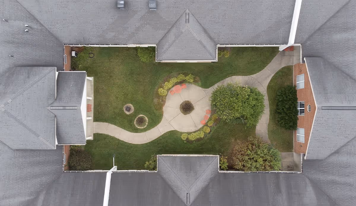 Aerial view of a courtyard garden enclosed by a building with a gray roof. The courtyard features a winding concrete pathway, green grass, several bushes, trees, and a few red chairs arranged around the pathway.