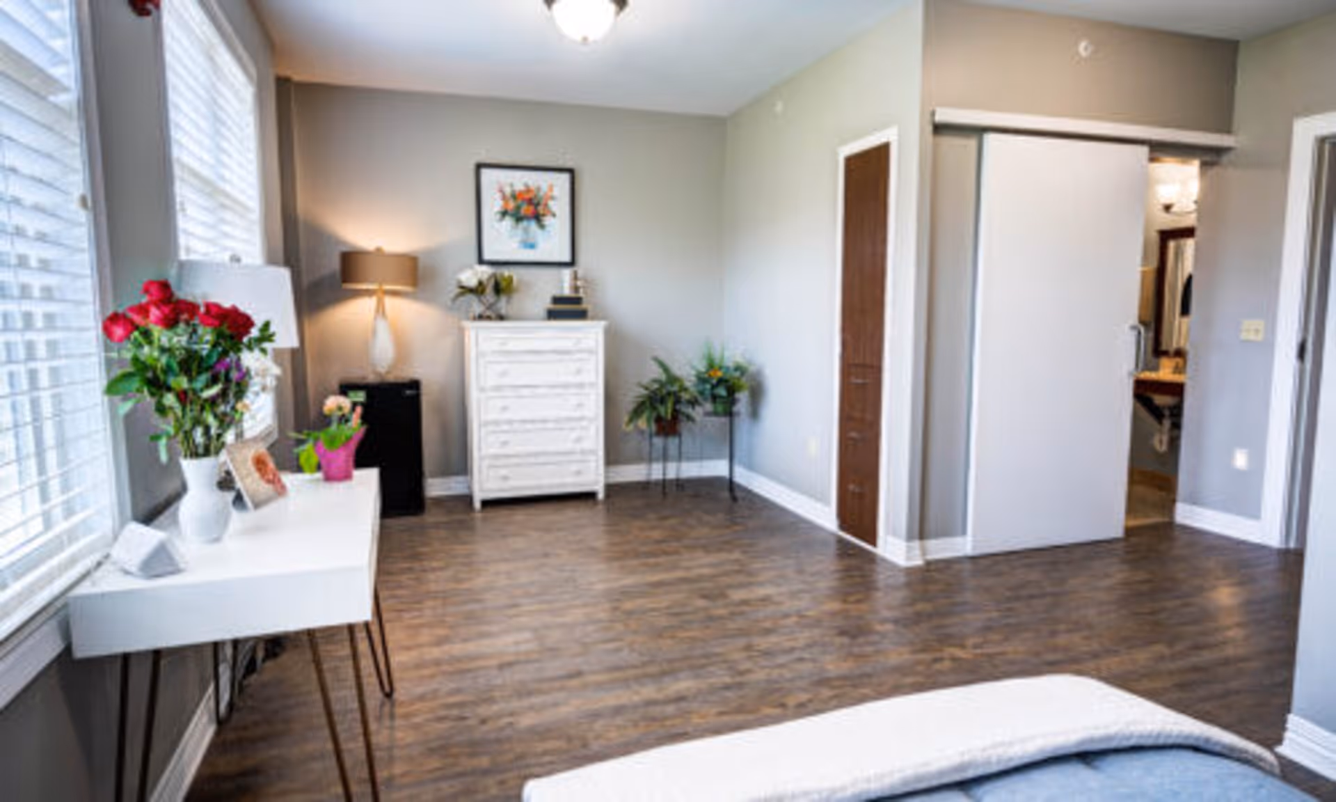 Bright bedroom with hardwood floors, a white dresser, a desk by large windows, decorative plants and a partially visible bed.