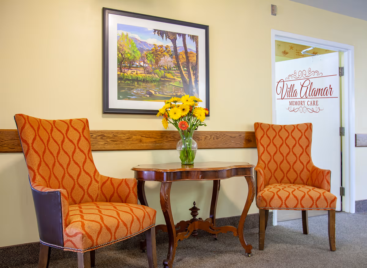 Two orange patterned armchairs with wooden legs are placed on either side of a small wooden table with curved legs. A glass vase with yellow and orange flowers sits on the table. Behind the chairs is a beige wall with a wooden chair rail and a framed painting of a landscape with trees and mountains. To the right, there is a white door with glass that has the text 'Villa Alamar Memory Care' written in red.