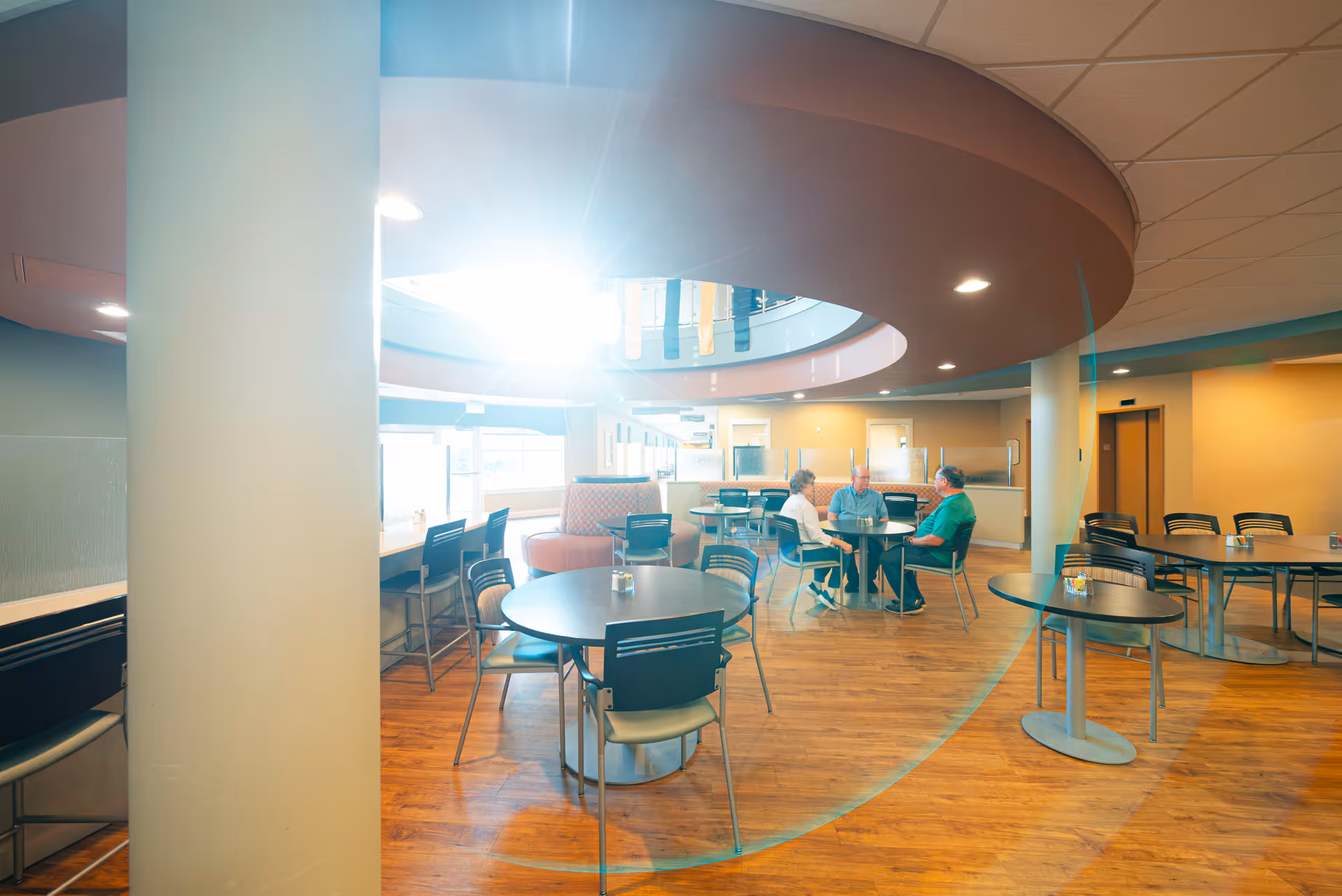 A bright, modern dining area with round tables and chairs arranged on a wooden floor. Three elderly people are seated at one of the tables engaged in conversation. The ceiling features a circular architectural design with recessed lighting, and large windows allow natural light to flood the space.