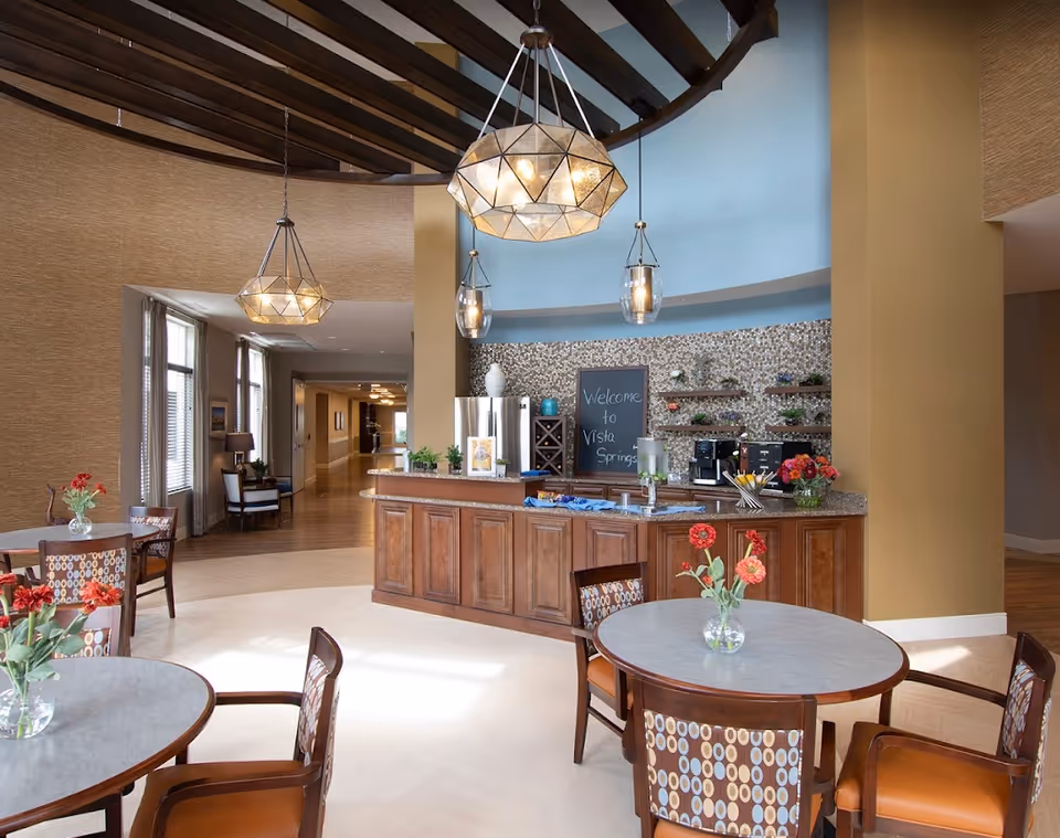 A bright and welcoming dining area in a senior living facility with round tables and chairs, each table decorated with a vase of red flowers. The room features a curved wooden counter with a granite countertop, a coffee machine, and a chalkboard sign that reads 'Welcome to Vista Springs'. The ceiling has exposed wooden beams and modern pendant lights hanging down. Large windows allow natural light to fill the space.