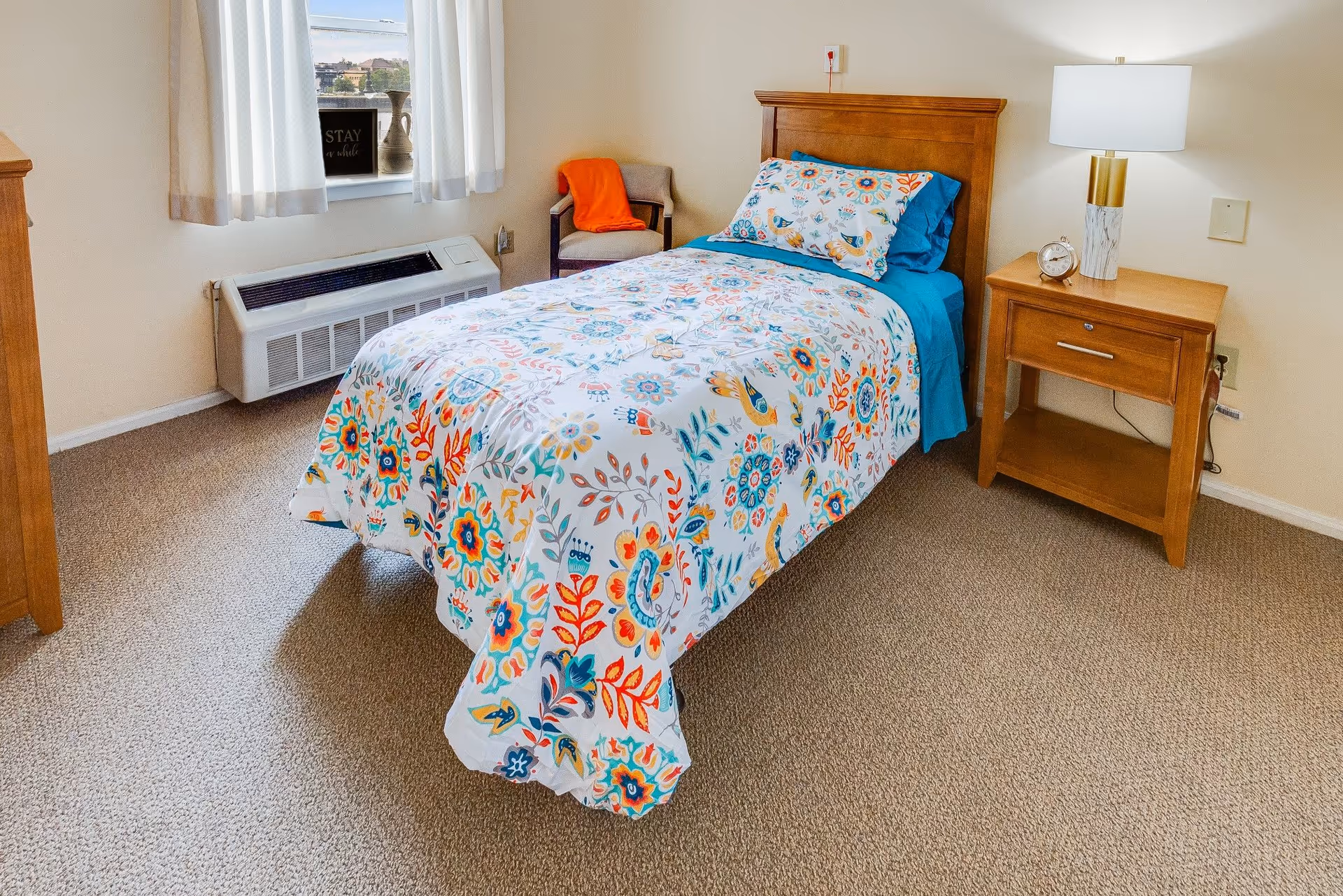 A neatly made single bed with a colorful floral and bird patterned bedspread and matching pillow in a small bedroom. Next to the bed is a wooden nightstand with a lamp and a small clock. There is a chair with an orange blanket draped over it in the corner near a window with white curtains. Below the window is a wall-mounted air conditioning unit.
