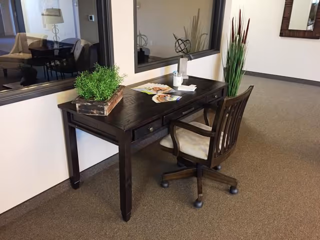 A dark wooden desk with a matching swivel chair on a carpeted floor. On the desk are a small potted plant, some magazines, and a cup holding pens. Behind the desk is a large window looking into another room, and to the right is a tall decorative plant. The setting appears to be a quiet interior space, possibly a hallway or common area in a facility.