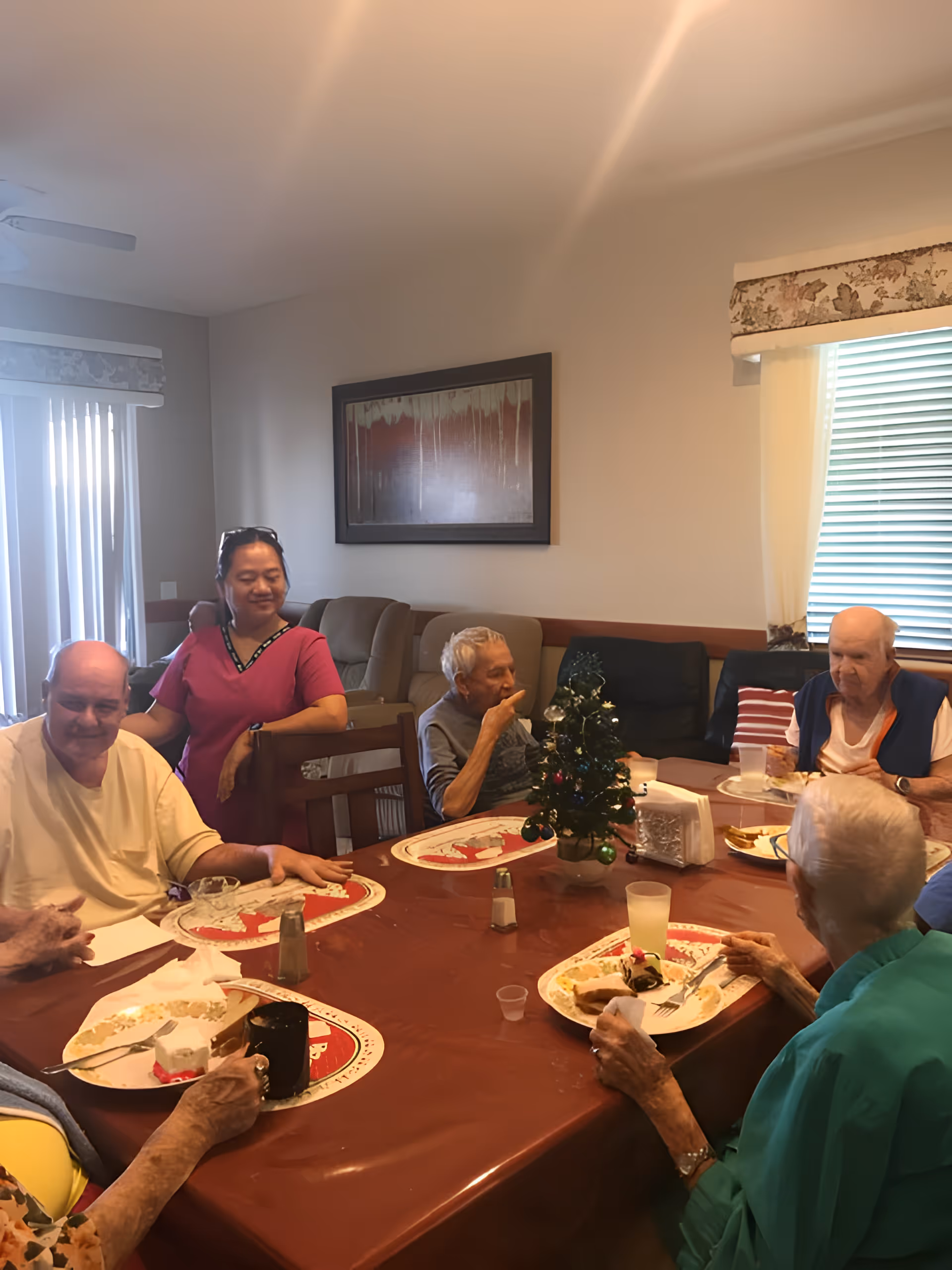 A group of elderly people sitting around a dining table with a small decorated Christmas tree in the center. A caregiver in a pink uniform stands behind one of the seated men, smiling. The room has beige walls, a window with blinds and curtains, and a framed abstract painting on the wall.