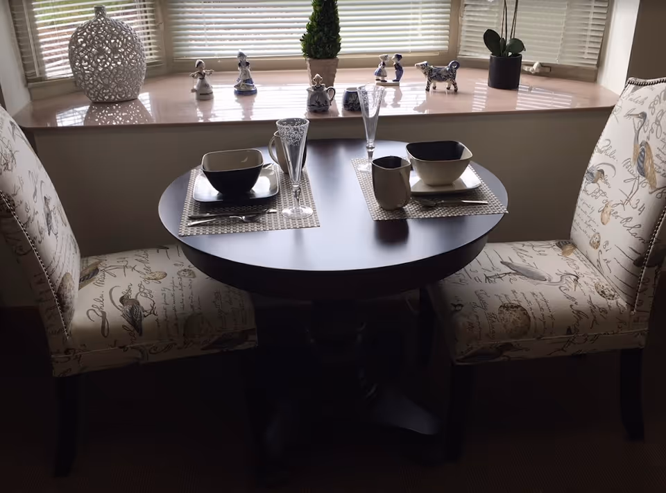 A small round dining table set for two with matching patterned chairs. The table has two place settings with bowls, plates, mugs, and tall glasses. Behind the table is a window sill with decorative figurines and a small potted plant.