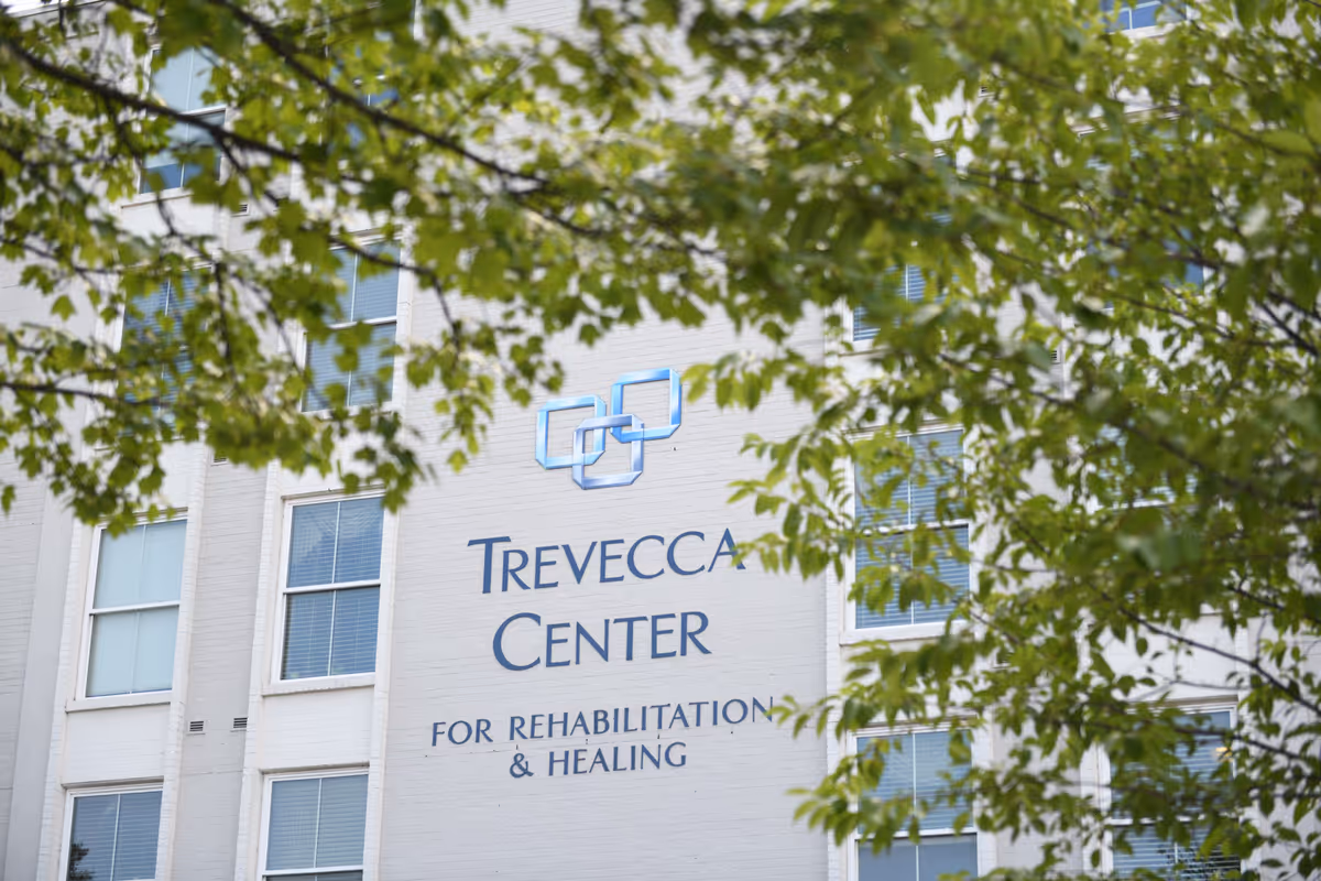 Exterior view of the Trevecca Center for Rehabilitation and Healing building partially obscured by green tree branches with leaves. The building is light-colored with several windows and the facility's name and logo are prominently displayed on the wall.