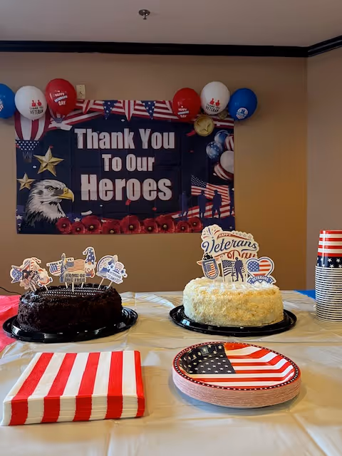 Table with two decorated cakes, American flag-themed plates and napkins set up in front of a 'Thank You To Our Heroes' banner and balloons.