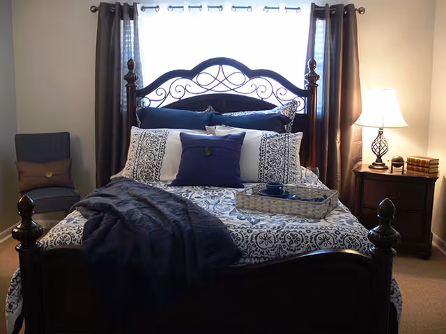 A neatly made bed with a dark wooden frame and decorative headboard, adorned with patterned white and blue bedding and multiple pillows. A dark blue throw blanket is draped over the bed. A wicker tray with a cup and saucer is placed on the bed. To the right, there is a wooden nightstand with a lamp and a small stack of books. A blue chair with a brown cushion is positioned to the left of the bed. Brown curtains frame a window behind the bed.