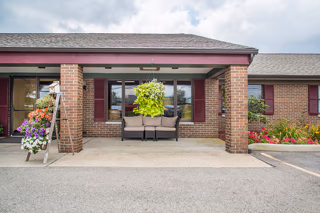 Front exterior view of a brick building with a covered entrance supported by brick columns. There is a bench with cushions under the covered area and a hanging plant above it. To the left, a wooden ladder holds several colorful flower pots. The sky is partly cloudy.