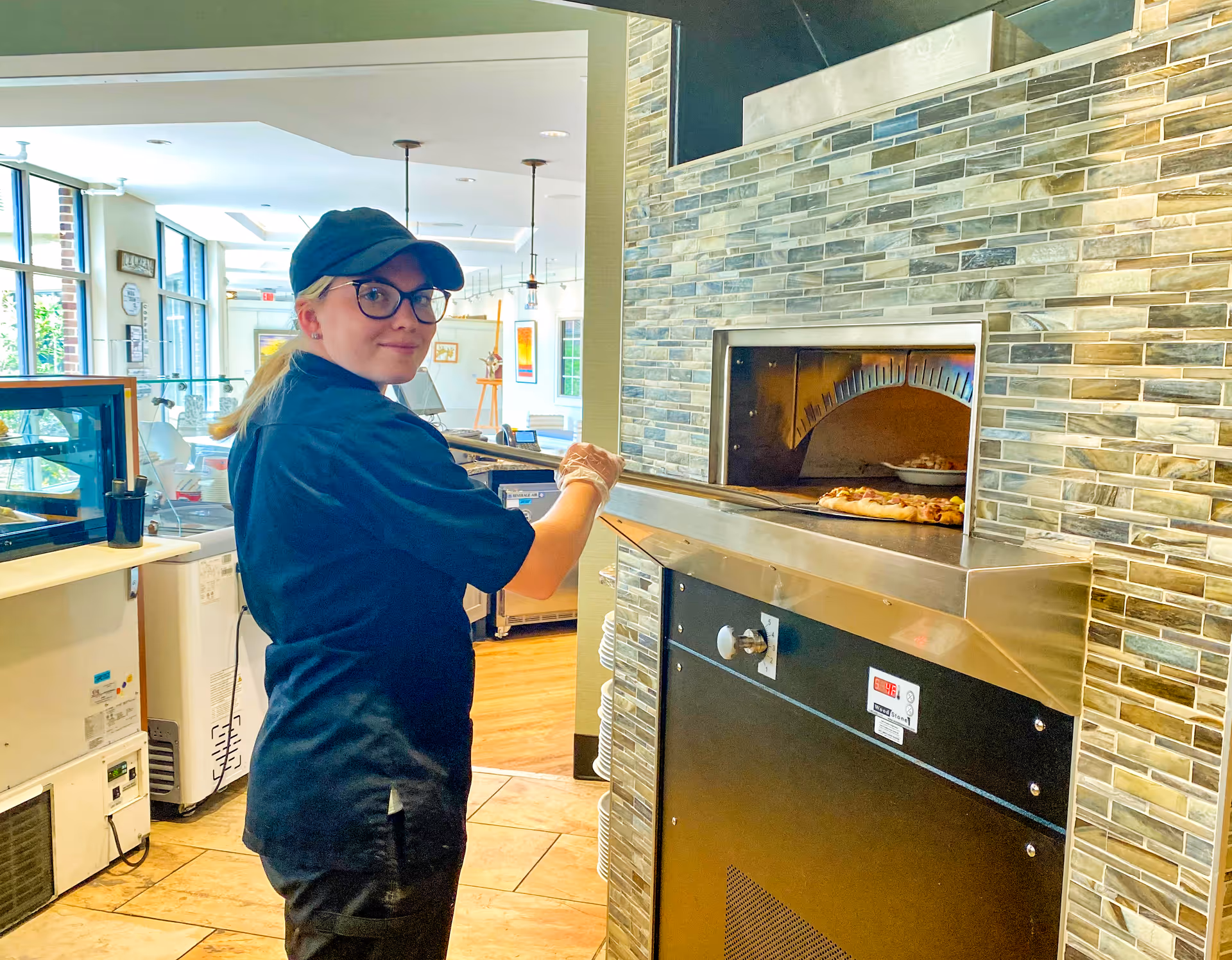 A woman wearing a black uniform, cap, and glasses is using a pizza peel to place or remove a pizza from a stone pizza oven in a bright kitchen area with large windows and tiled floors.