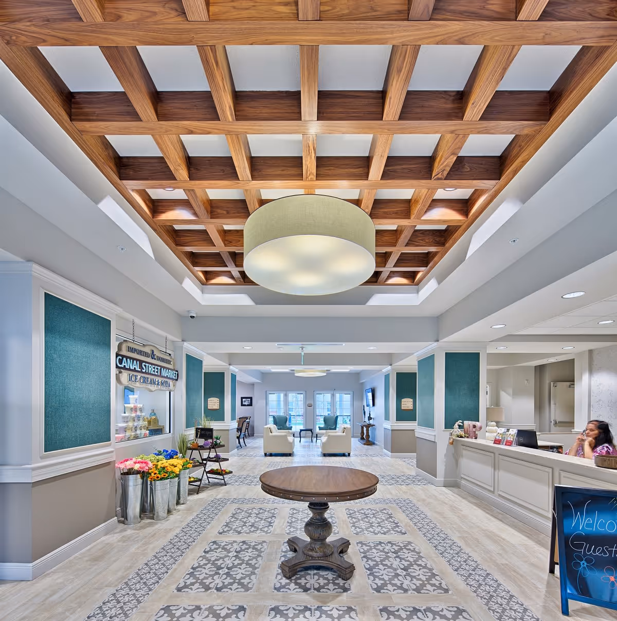 Interior view of a senior living facility lobby with a decorative wooden grid ceiling and a large round light fixture. The floor has patterned tiles in the center with a round wooden table. To the left, there are metal buckets with colorful flowers and a sign for Canal Street Market offering ice cream and soda. To the right, a reception desk with a woman sitting behind it and a chalkboard sign welcoming guests. In the background, there are seating areas with armchairs and large windows letting in natural light.
