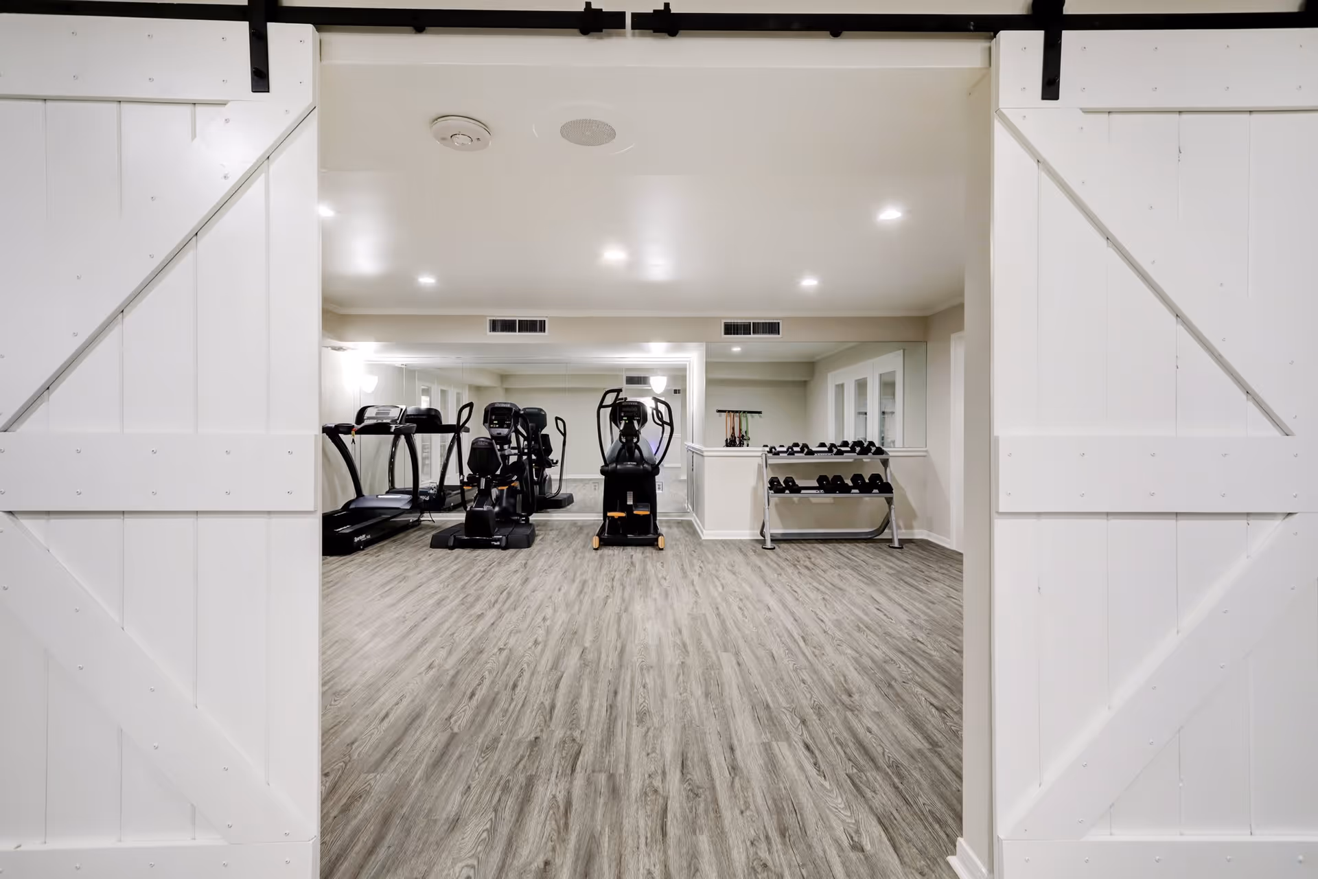 View through white barn-style sliding doors into a fitness room with wood-look flooring, exercise machines including treadmills and ellipticals, a rack of dumbbells, and a large mirror on the back wall.