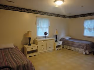 A simple bedroom with two single beds placed against opposite walls, each with a pillow and striped bedspread. Between the beds is a white dresser with six drawers and two matching nightstands, each holding a lamp. Two windows with sheer white curtains allow natural light into the room. The walls are light-colored with a decorative border near the ceiling.