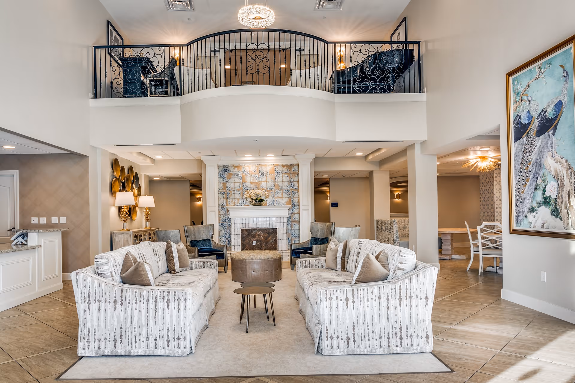 A spacious and elegant senior living facility common area featuring two patterned sofas with multiple throw pillows facing each other across a small round coffee table. Behind the seating area is a decorative fireplace with intricate tile work. Two armchairs with blue cushions flank the fireplace. Above, there is a balcony with ornate black railing and two chairs. The room has light-colored walls, tiled floors, and a large framed painting of peacocks on the right wall. To the left is a reception desk with lamps and decorative wall art.