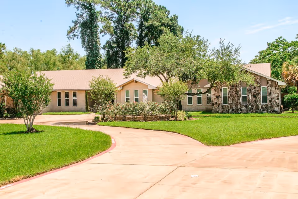 Exterior view of a single-story assisted living facility building with a stone and beige facade, surrounded by green grass, trees, and bushes under a clear blue sky. A curved concrete driveway leads up to the building.