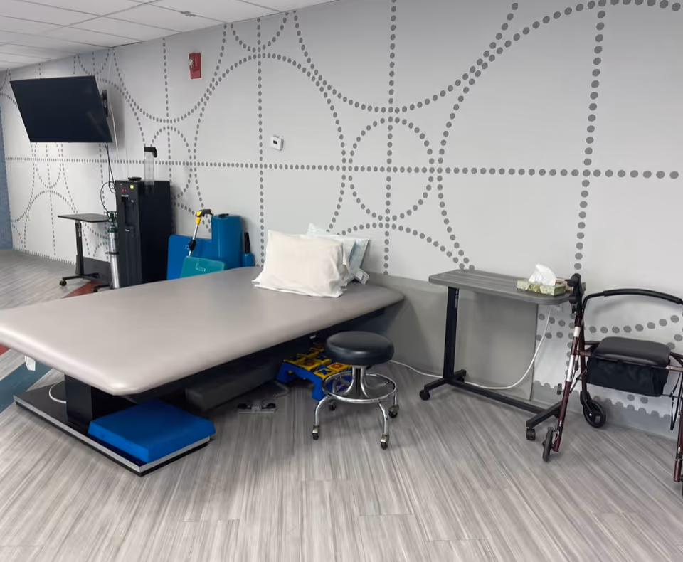 A therapy or treatment room with a large padded table, a small rolling stool, a walker, a small table with tissues, and a wall-mounted TV. The wall has a decorative pattern of dotted circles and lines, and the floor is covered with light-colored wood-like flooring.