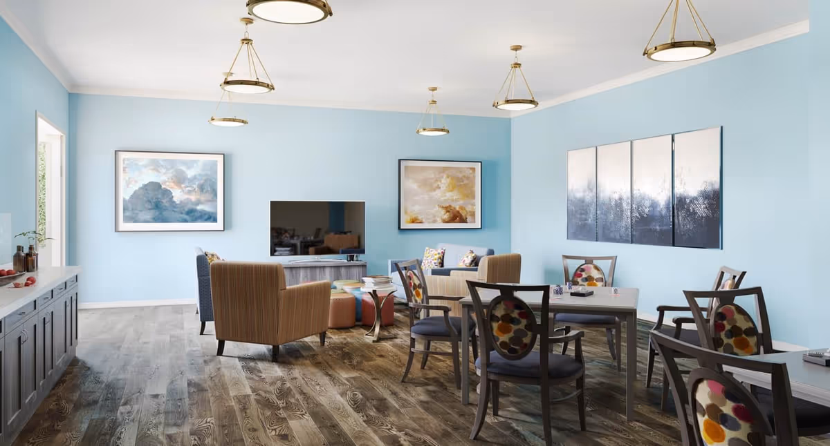 Communal dining and lounge area with dining tables and chairs, armchairs around a TV, and light blue walls.