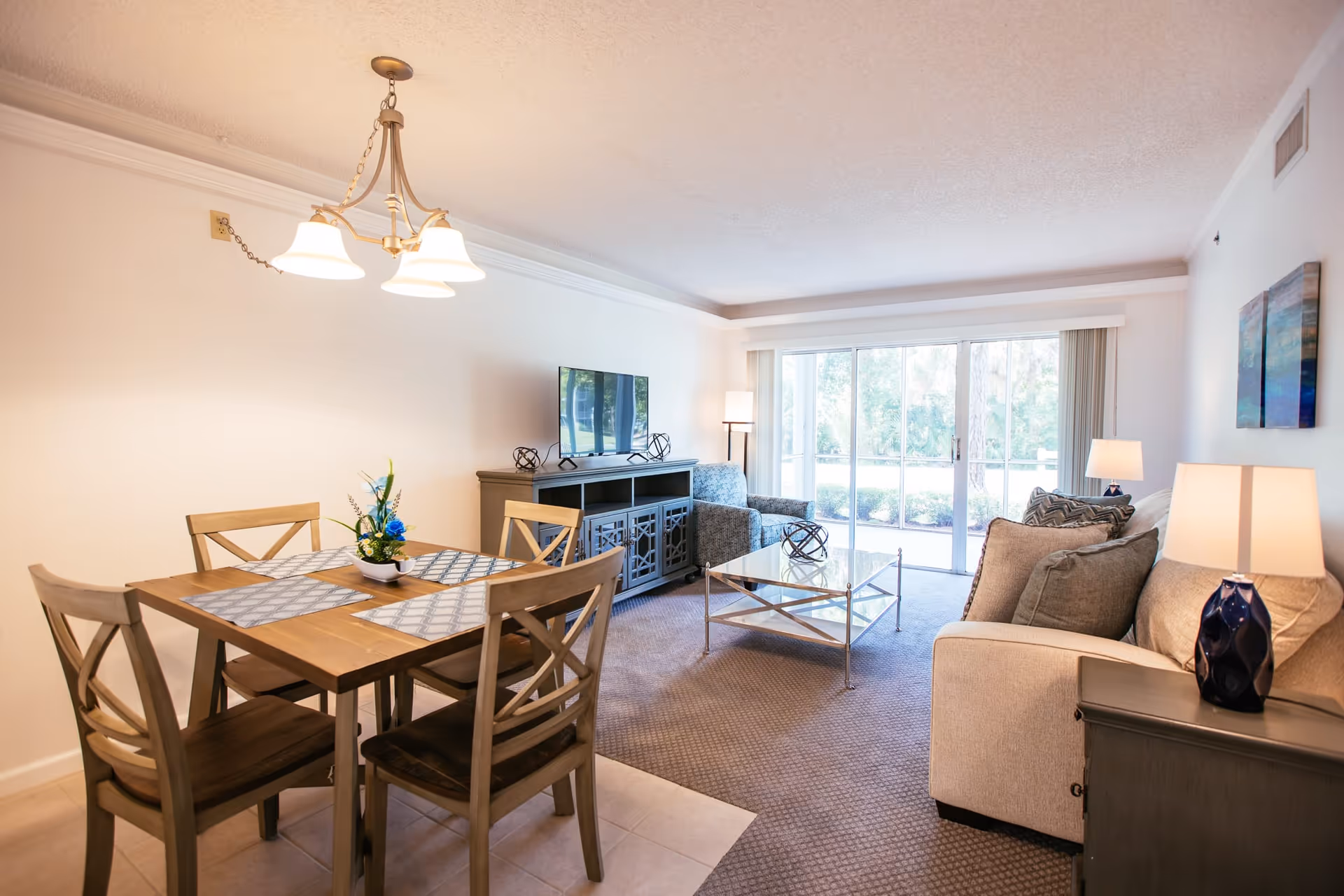 A bright and cozy living and dining area in a senior living facility. The dining area features a wooden table with four chairs and a small flower arrangement centerpiece. The living area has a beige sofa with cushions, a glass coffee table, an armchair, a TV on a dark wooden stand, and two table lamps. Large sliding glass doors let in natural light and offer a view of greenery outside.